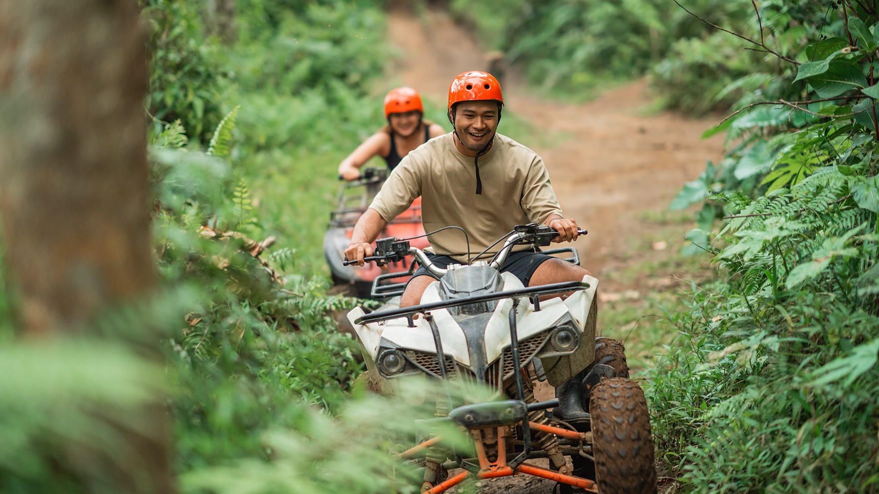 Riders in orange helmets following a guide along a narrow jungle track on quad bikes