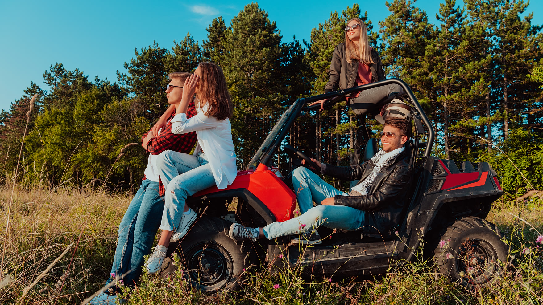 Four people sitting on and around a red four-seater roll-caged off-road buggy at the edge of a hillside track