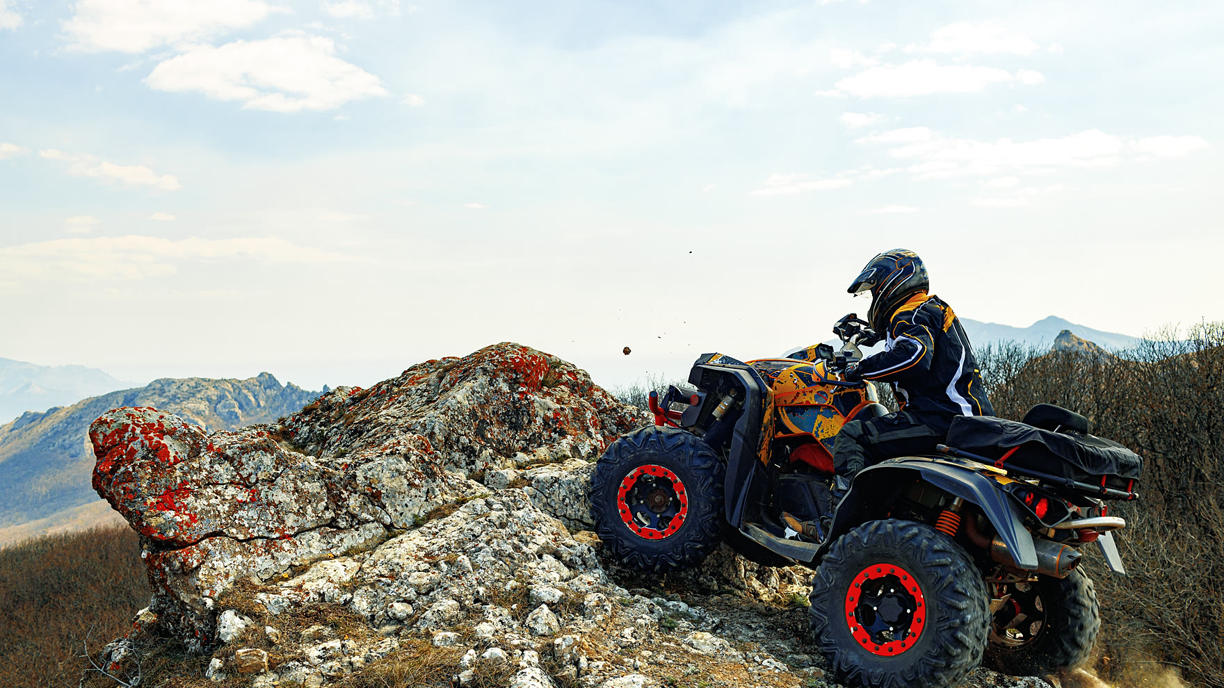 Rider in a full-face helmet and body armour on a quad bike at the edge of a rocky ridge above forested hills